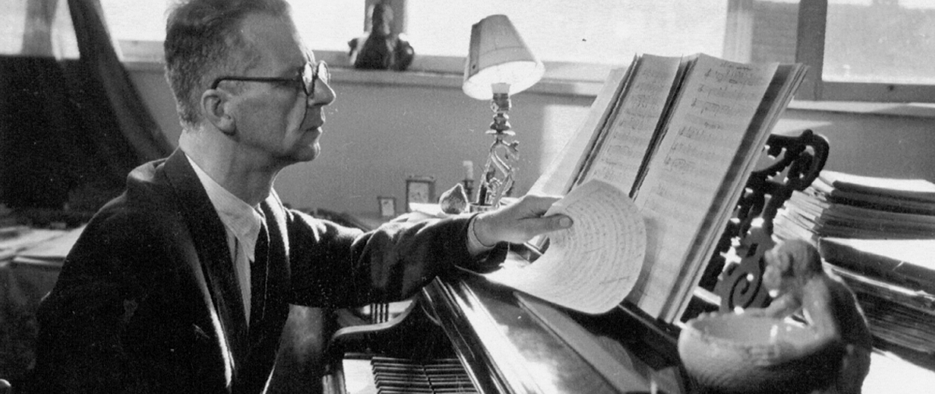 Zdjęcie przedstawia Zygmunta Mycielskiego siedzącego przy fortepianie, patrzącego na nuty stojące na pulpicie. W lewej dłoni trzyma nuty. Na fortepianie za pulpitem widoczny stos dokumentów oraz miseczka z figurką małpki. W tle lampka oraz okna. Całość zdjęcia czarno-biała.
