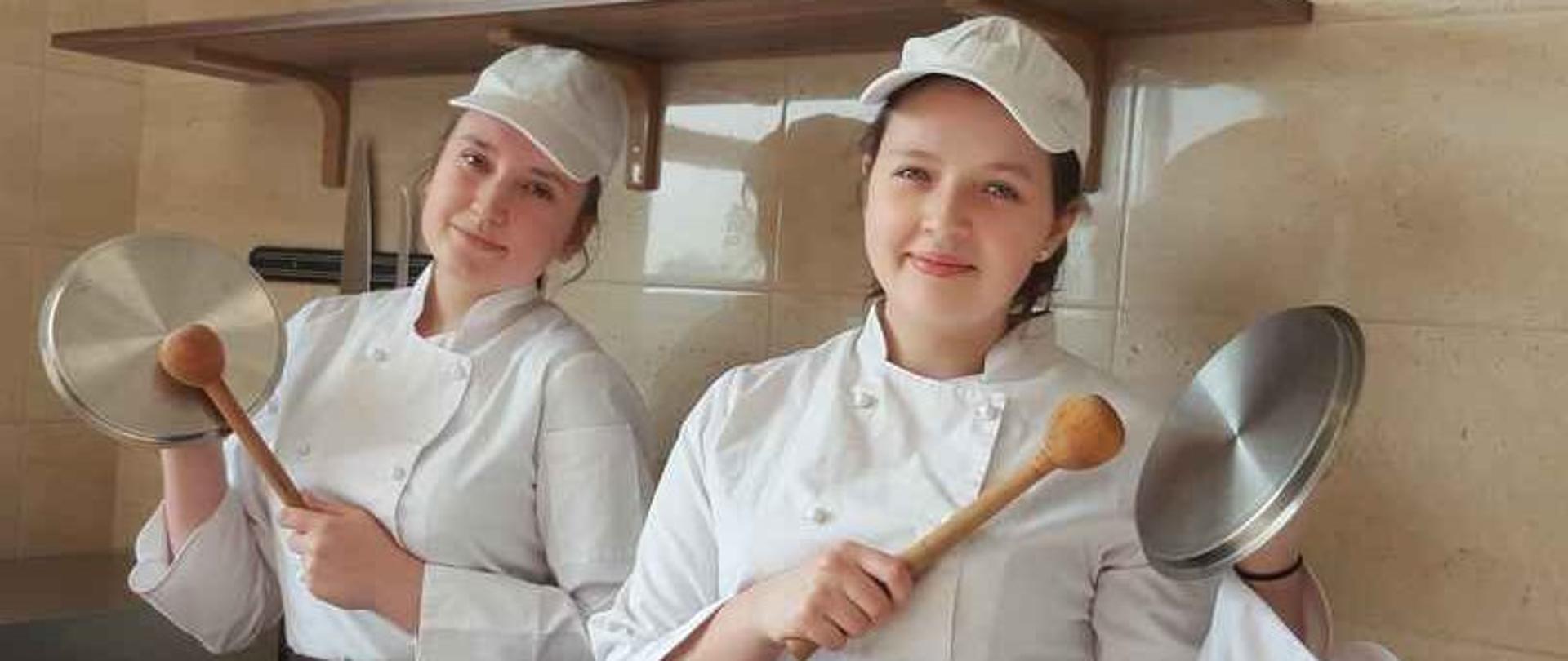 W kuchni stoją dwie dziewczyny. Każda z nich trzyma w ręku pokrywkę oraz pałkę do ucierania ciasta.