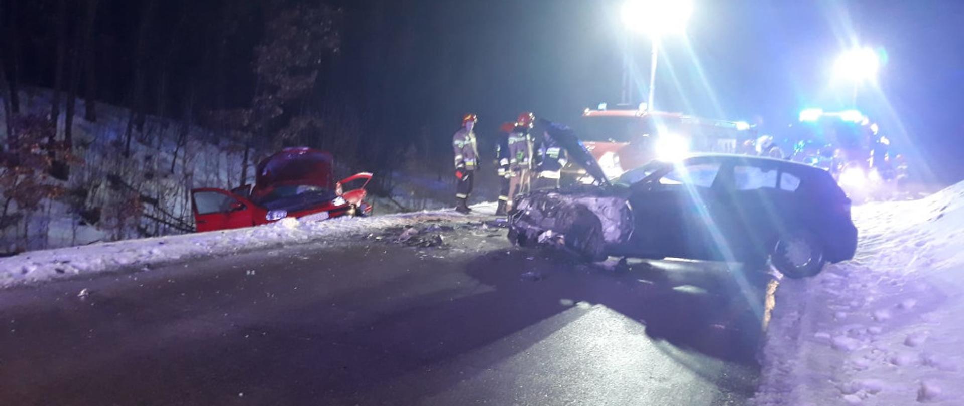 Zdjęcie przedstawia dwa uszkodzone samochody osobowe. Jeden z nich w kolorze czerwonym znajduje się w rowie z otwartą maską. Drugi stoi poprzecznie do osi jezdni na jednym pasie ruchu. Na drugim planie znajduje się kilku strażaków w ubraniach bojowych z założonymi czerwonymi hełmami na głowach. Za nimi znajdują się dwa samochody ratowniczo-gaśnicze z włączonymi włączonymi błyskowymi światłami alarmowymi w kolorze niebieskim oraz masztami oświetleniowymi. Zdjęcie zrobione w porze nocnej. Na poboczu zalega biała warstwa śniegu.