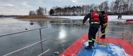 Zdjęcie przedstawia strażaków stojących na pomoście, którzy rzucają rzutkę do osoby pod którą się zarwał lód. 