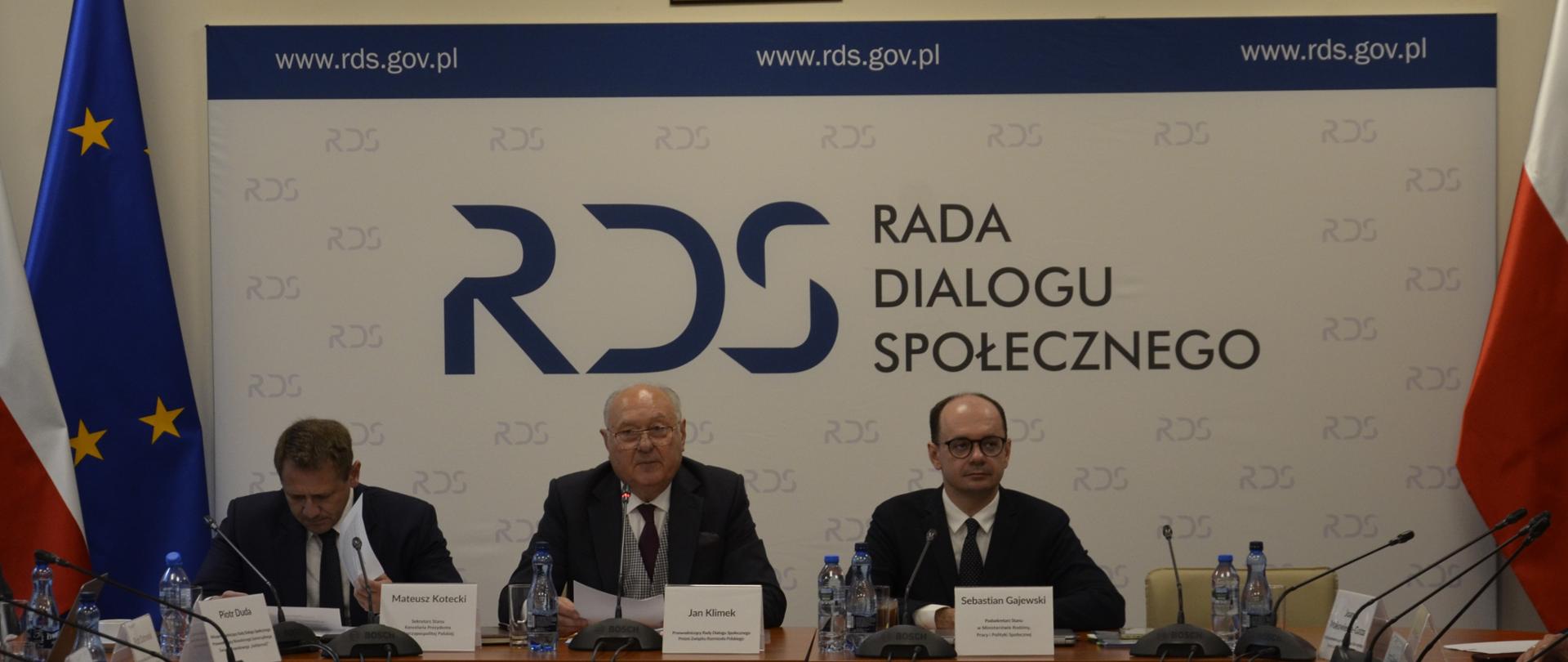 Na zdjęciu widnieje stół prezydialny za którym siedzą: Pan Mateusz Kotecki – Sekretarz Stanu w Kancelarii Prezydenta RP; Pan Jan Klimek, Przewodniczący Rady Dialogu Społecznego oraz Pan Sebastian Gajewski, Podsekretarz Stanu w Ministerstwie Rodziny, Pracy i Polityki Społecznej. W tle znajduje się baner z napisem Rada Dialogu Społecznego. Po lewej stronie baneru stoją flagi Polski oraz UE. Po prawej stronie baneru znajduje się flaga Polski. Nad banerem znajduje się Godło Polski.