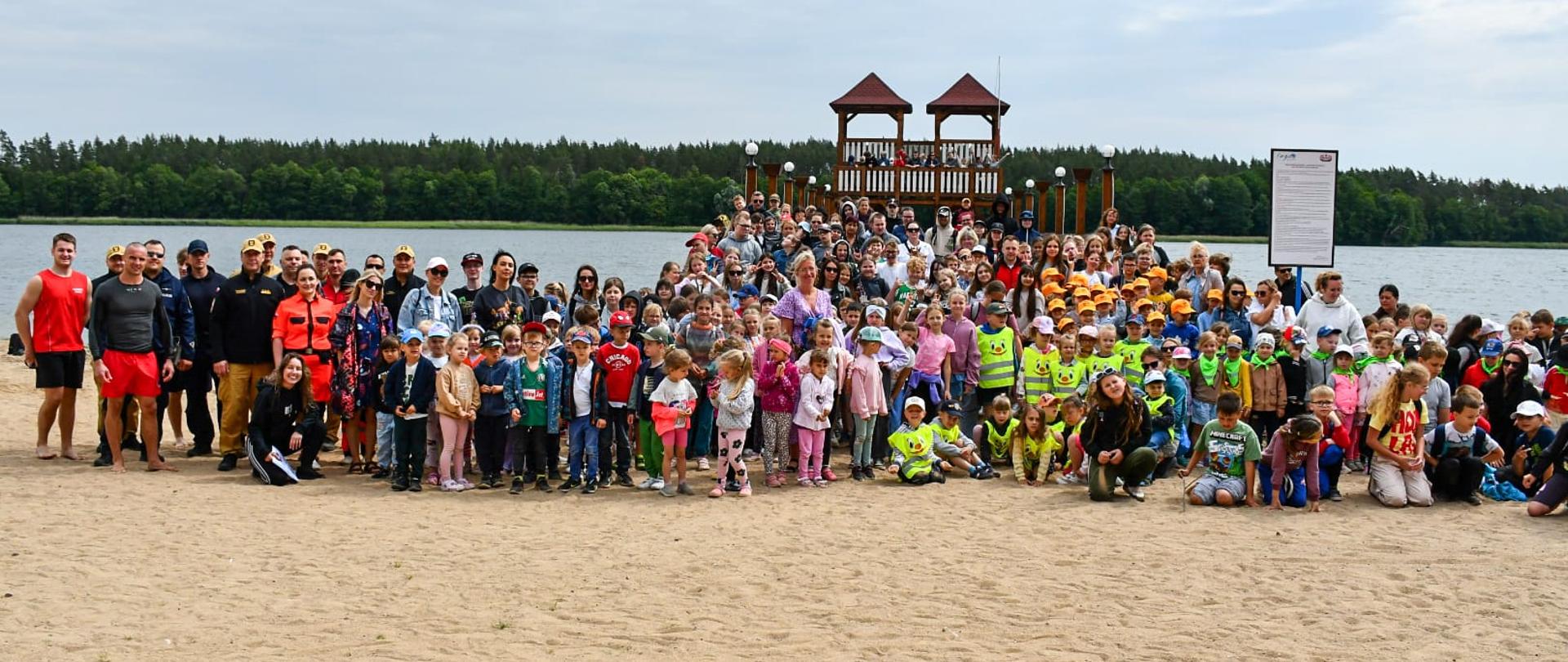 Grupa uczestników pokazu stojąca na plaży. W tle widoczne są drewniane wieże, które są częścią molo, jezioro oraz las.