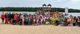 Grupa uczestników pokazu stojąca na plaży. W tle widoczne są drewniane wieże, które są częścią molo, jezioro oraz las.