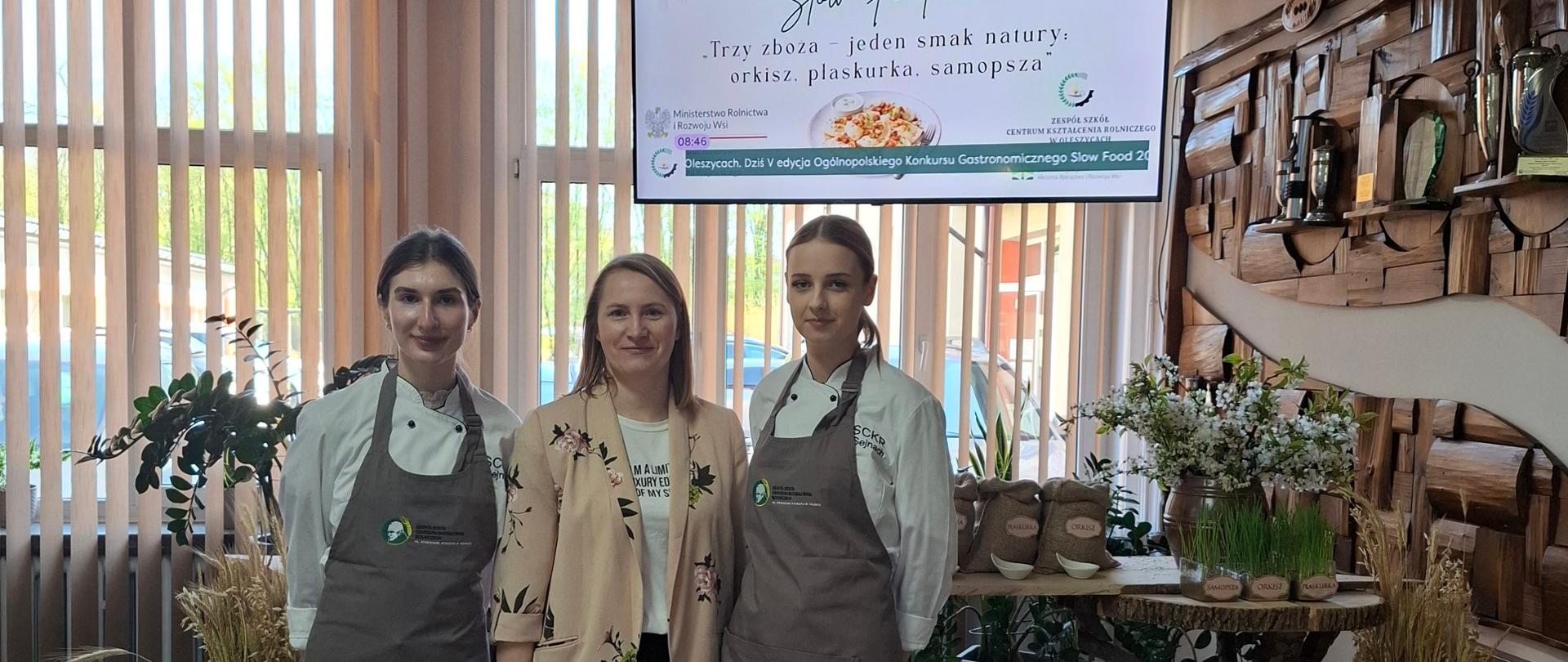 Dwie uczennice w stroju kucharza i nauczycielka stoją na tle stołu, na którym wyeksponowano woreczki ze starymi odmianami zbóż.