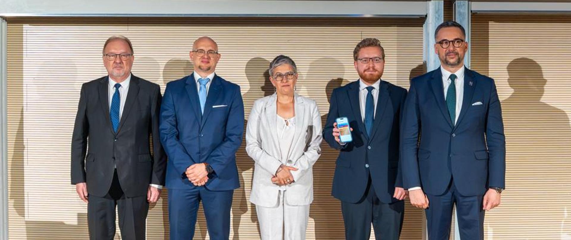 Przed ścianą z cienkich poziomych listewek stoi pięć osób, dwóch mężczyzn w garniturach, pośrodku wiceminister Mrówczyńska w jasnoszarej marynarce, dwóch mężczyzn w garniturach, pierwszy z prawej minister Kulasek.
