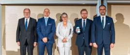 Przed ścianą z cienkich poziomych listewek stoi pięć osób, dwóch mężczyzn w garniturach, pośrodku wiceminister Mrówczyńska w jasnoszarej marynarce, dwóch mężczyzn w garniturach, pierwszy z prawej minister Kulasek.