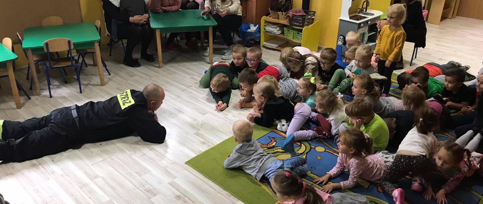 W sali przedszkola na podłodze strażak leży na brzuchu, przed nim leży kilkoro dzieci, część siedzi w tle przy stoliku siedzą opiekunki. Strażak przedstawia bezpieczne zachowanie w dymie.