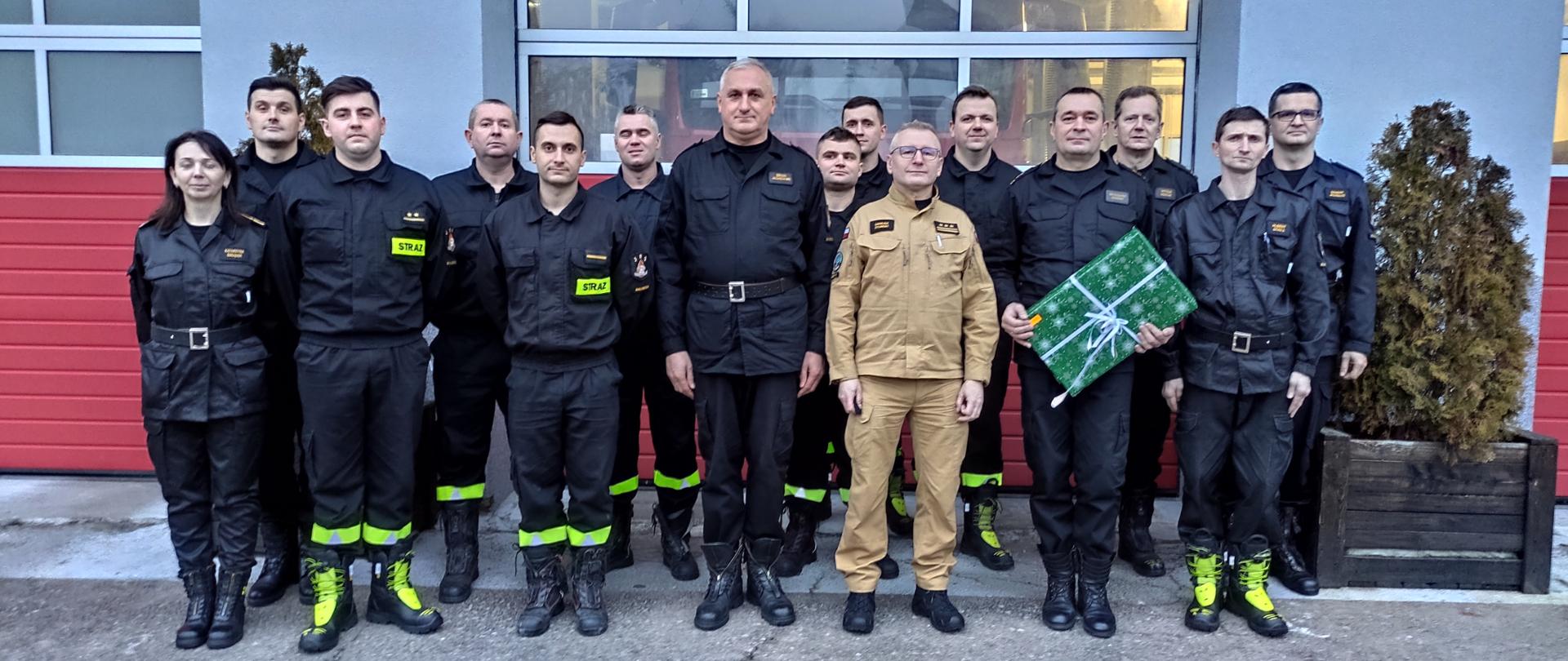 Strażacy z Komendy Powiatowej Państwowej Straży Pożarnej w Białobrzegach stoją przed garażami. Na czele stoi Zastępca Mazowieckiego Komendanta Wojewódzkiego Państwowej Straży Pożarnej. Komendant Powiatowy Państwowej Straży Pożarnej w Białobrzegach trzyma w rękach zapakowany prezent.