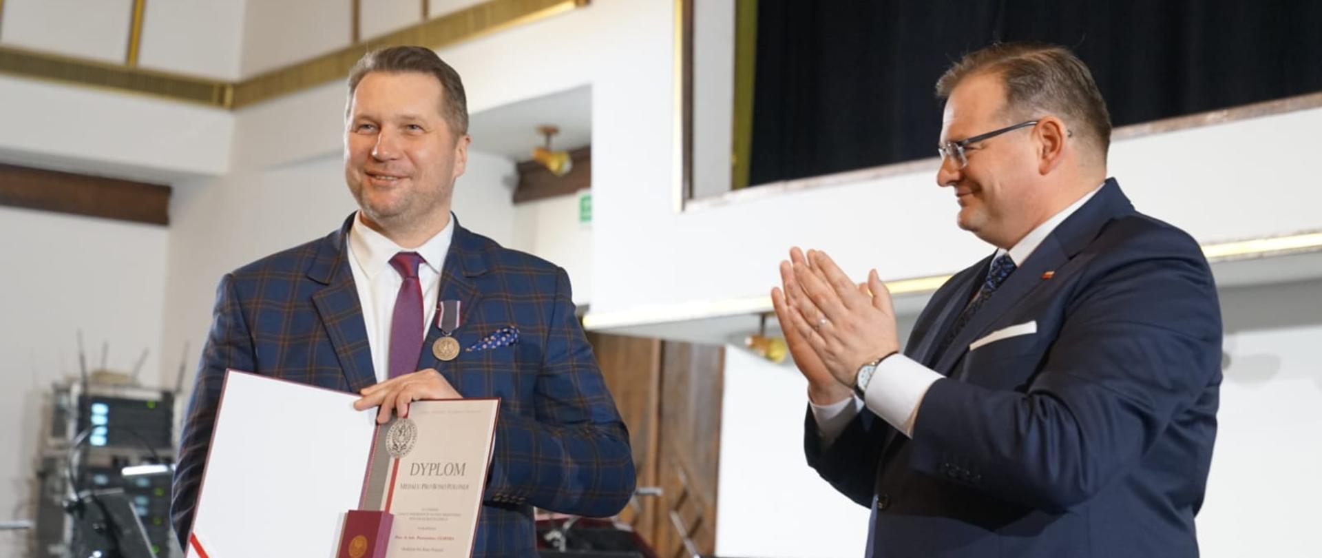 Uśmiechnięty minister Czarnek stoi na tle białej ściany i pokazuje otwarty dokument, obok niego mężczyzna w szarym garniturze bije brawo.