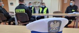 Zdjęcie na pierwszym planie przedstawia białą czapkę garnizonową policji z jedną gwiazdką. W tle funkcjonariusze z Wydziału Ruchu Drogowego Komendy Powiatowej Policji w Jędrzejowie, którzy prowadzą prelekcje z zakresu zasad bhp i przepisów dotyczących bezpieczeństwa ruchu drogowego. Na świetlicy przy stolikach siedzą także strażacy w maseczkach zasłaniających usta i nos. 