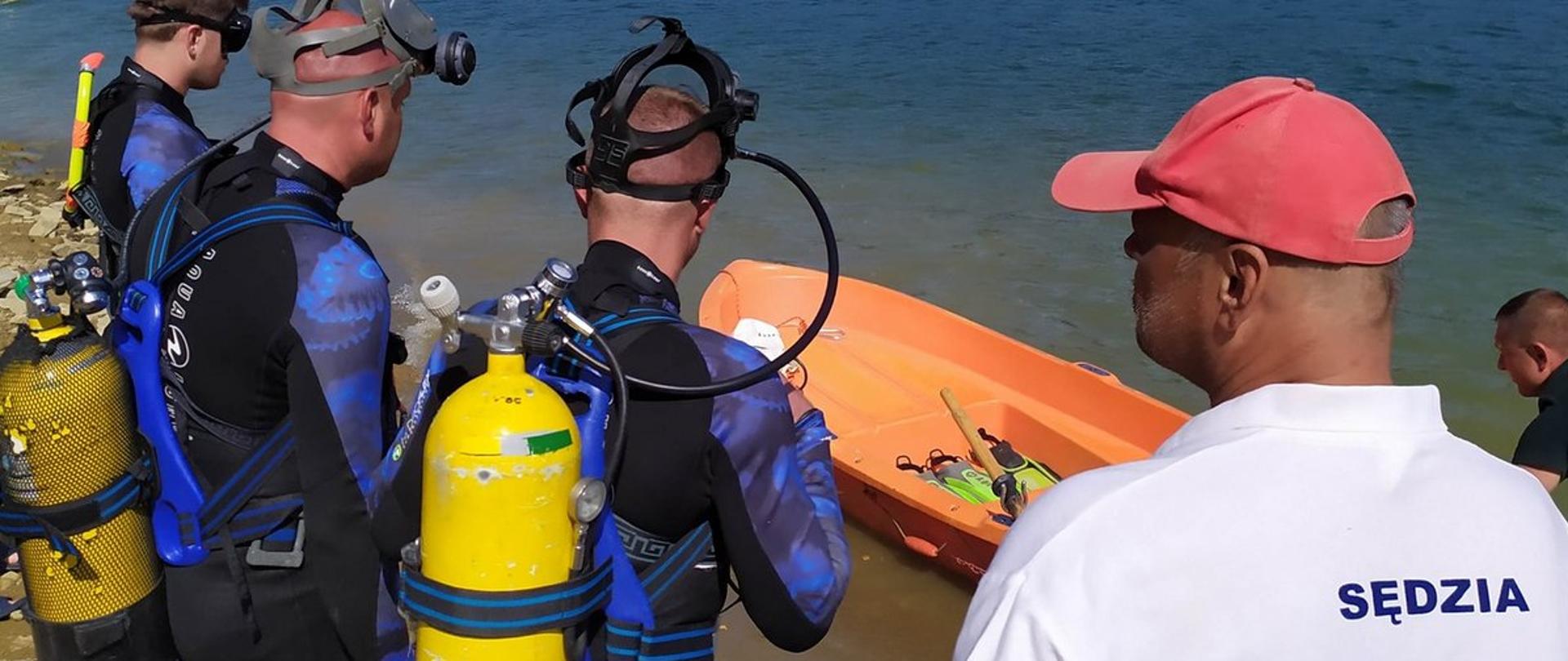 Na zdjęciu widać trzech płetwonurków obróconych tyłem. Po prawej stronie zdjęcia mężczyzna w koszulce z napisem sędzia na plecach. Przed mężczyznami zbiornik wodny na którym jest łódka. 
