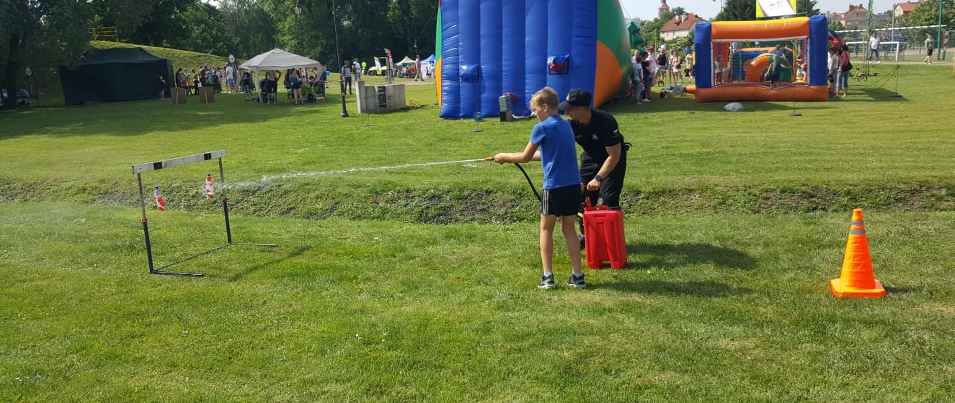 Dziecko leje wodę na butelki przy pomocy hydronetki. Strażak mu pomaga. Za nimi w oddali widać namioty dmuchane do zabawy dla dzieci oraz widać namioty stelażowe przy których stoją ludzie. 