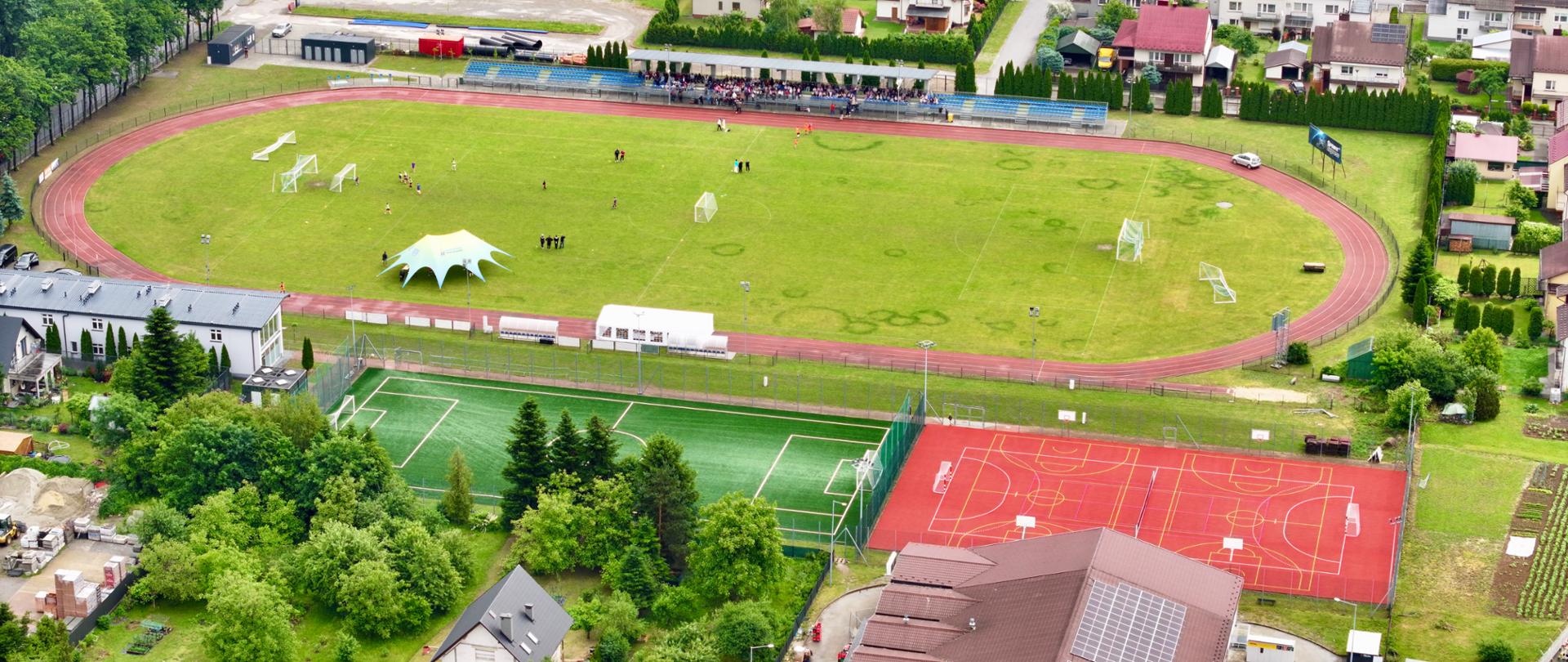 Widok z lotu ptaka na boisko piłkarskie. Obok niego Orlik i kort tenisowy. Wokół zabudowania.
