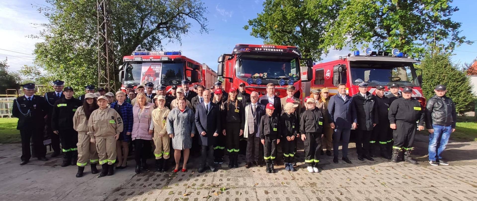 Na zdjęciu stoją uczestnicy spotkania, strażacy oraz zaproszeni goście. Za nimi trzy samochody strażackie. Słonecznie.