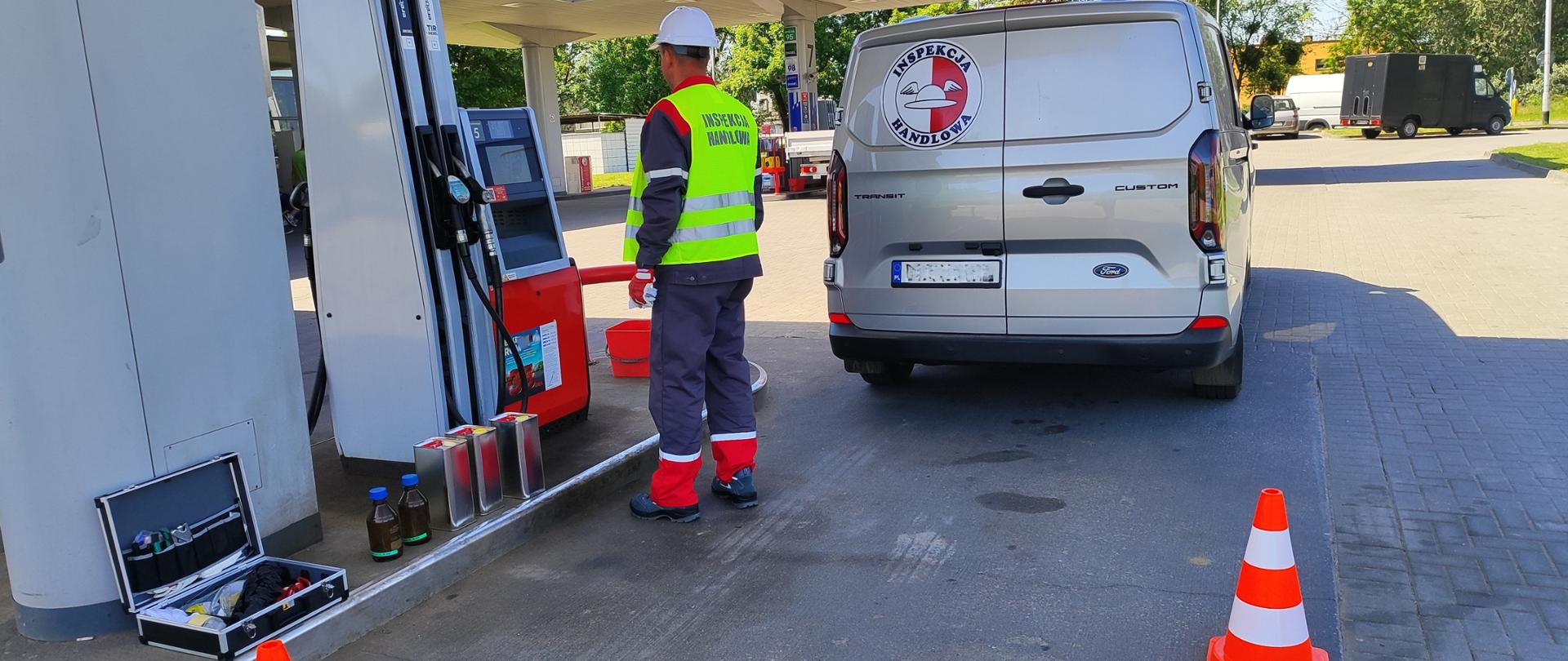 Pracownik Inspekcji Handlowej w trakcie czynności kontrolnych na stacji paliw stoi przed dystrybutorem. Pod dystrybutorem stoją 3 metalowe pojemniki na próby i 2 szklane butelki na próby oraz walizka ze sprzętem. Obszar pracy jest odgrodzony pachołkami, w tle samochód Inspekcji Handlowej do przewożenia paliw i infrastruktura stacji. 