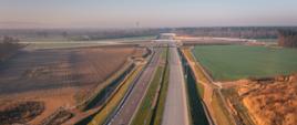 Zdjęcie przedstawia widok z lotu ptaka na wielopasmową autostradę biegnącą przez tereny wiejskie. Po obu stronach drogi rozciągają się pola uprawne, zielone łąki i kępy drzew. W oddali widoczny jest wiadukt lub most, a sama droga wygląda na nowoczesną i dobrze utrzymaną. Nad krajobrazem unosi się lekka mgiełka, a niebo pozostaje bezchmurne, co podkreśla spokojny charakter otoczenia.