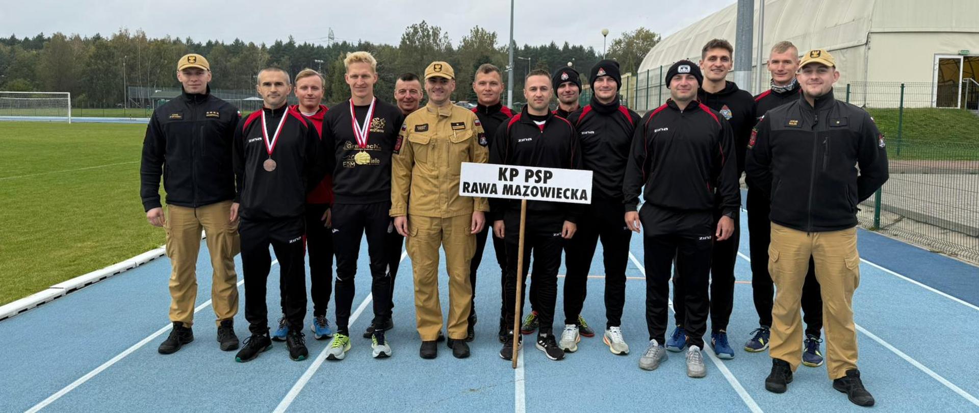 Zdjęcie przedstawia reprezentacje Komendy Powiatowej PSP w Rawie Mazowieckiej, która wzięła udział w XXIII Mistrzostwach Województwa Łódzkiego Strażaków PSP w lekkoatletyce. Na niebieskiej bieżni stoi 14 mężczyzn, stojących w rzędzie. Trzech z nich ubranych jest mundur służbowy strażacki, a reszta ubrana jest w czarno - czerwone dresy sportowe. Na środku stoi biała tabliczka z czarnym napisem KP PSP Rawa Mazowiecka.