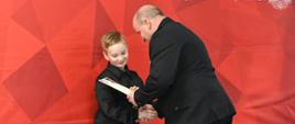 
Mężczyzna w marynarce ściska dłoń jednego z chłopców podczas uroczystego wręczenia nagród. Chłopcy stoją w rzędzie na tle czerwonego baneru z napisem „Państwowa Straż Pożarna” i symbolem orła, ubrani głównie na czarno; część z nich trzyma dyplomy oraz torby z upominkami. W tle widoczni są strażacy w mundurach, obserwujący ceremonię.
