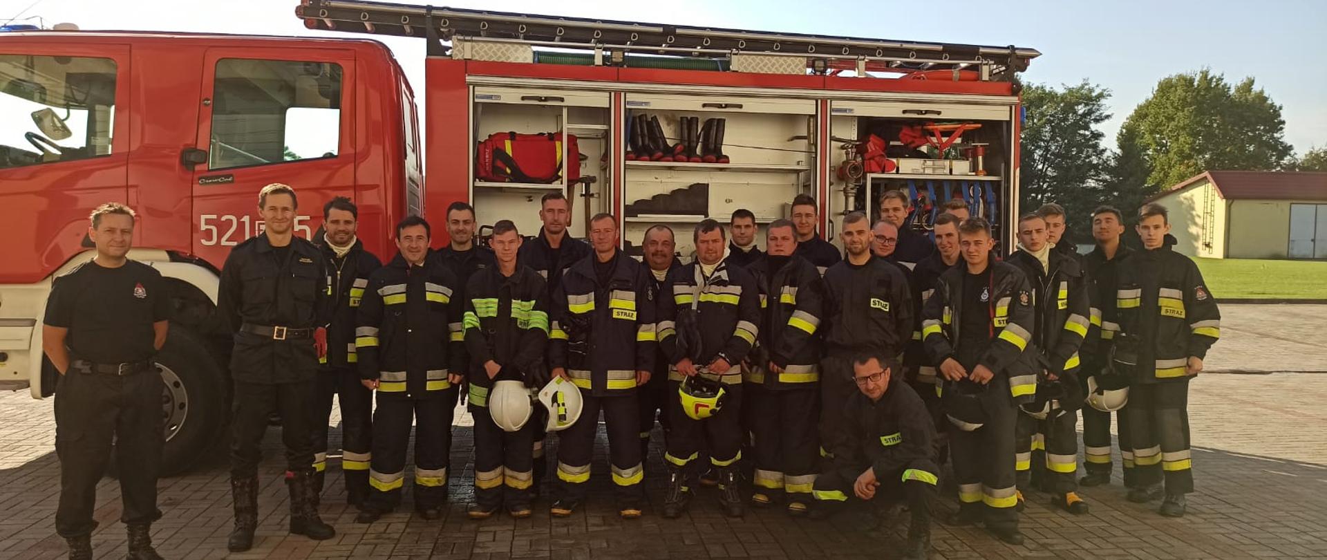 W dwuszeregu około 22 mężczyzn, druhów OSP, w strojach bojowych. Bezpośrednio za nimi stoi czerwony samochód gaśniczy z otwartym bokiem, szafkami na sprzęt.