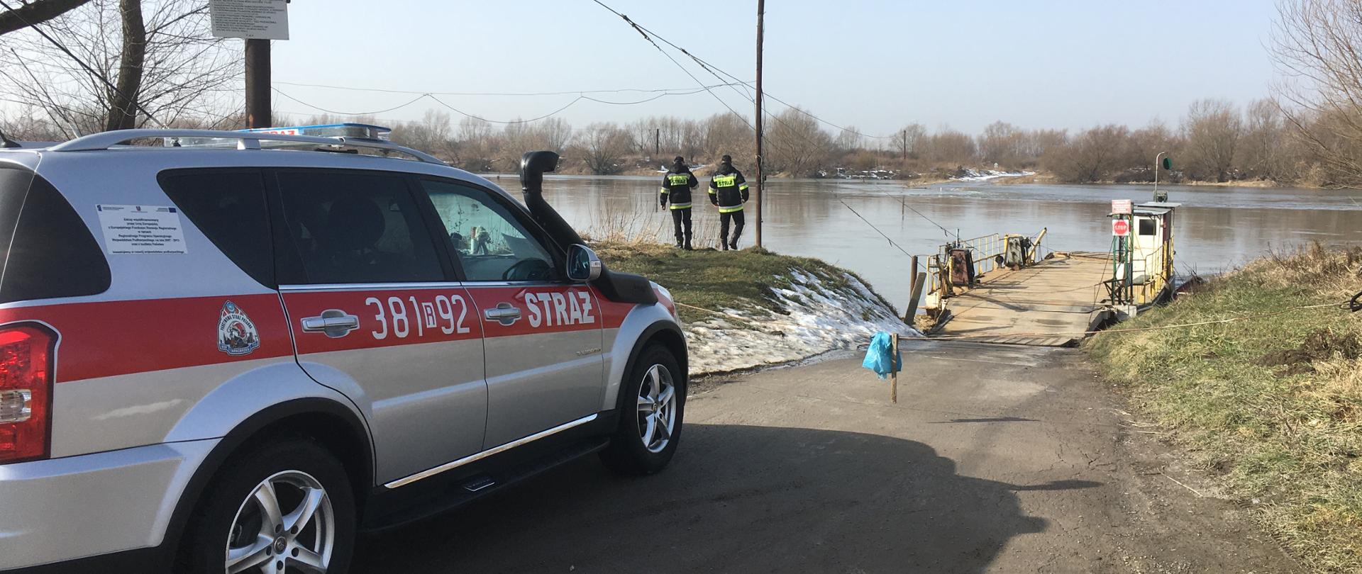Mały samochód strażacki stojący na drodze prowadzącej na prom. Nad samym brzegiem rzeki stoi dwóch strażaków droga prowadzi do promu, który jest zacumowany na rzece, ale wjazd na niego jest zablokowany.