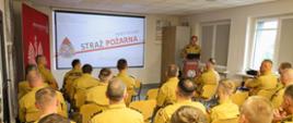 Grupa strażaków Państwowej Straży Pożarnej uczestniczy w podsumowaniu ćwiczeń w sali wykładowej. Prelegent stoi obok ekranu, na którym wyświetlany jest slajd prezentacji, a na podium obok laptopa widnieje logo PSP. Uczestnicy ubrani są w mundury służbowe z emblematami PSP i siedzą przodem do ekranu. W tle widoczne są plakaty informacyjne i tablice edukacyjne, co podkreśla formalny i edukacyjny charakter wydarzenia.