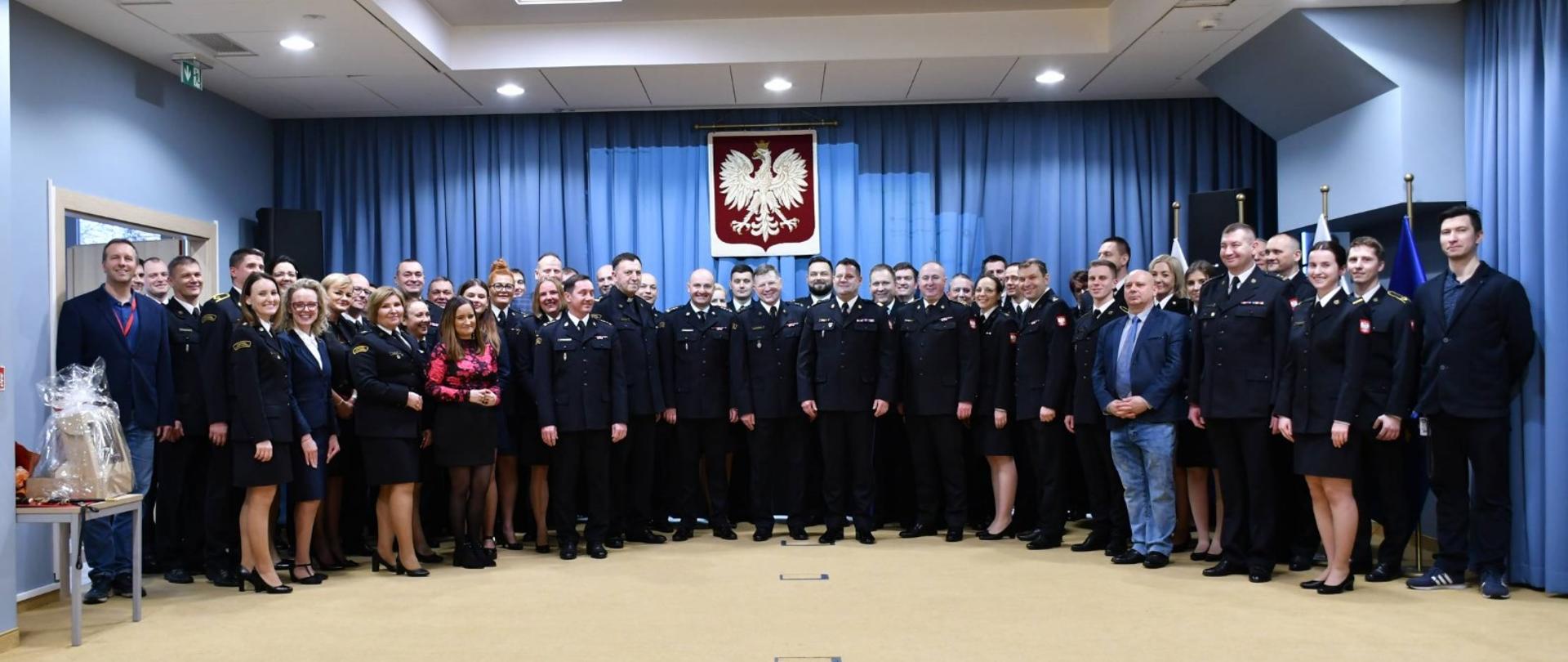 Zdjęcie grupowe wewnątrz pomieszczenia na tle niebieskiej ściany z godłem Polski. 