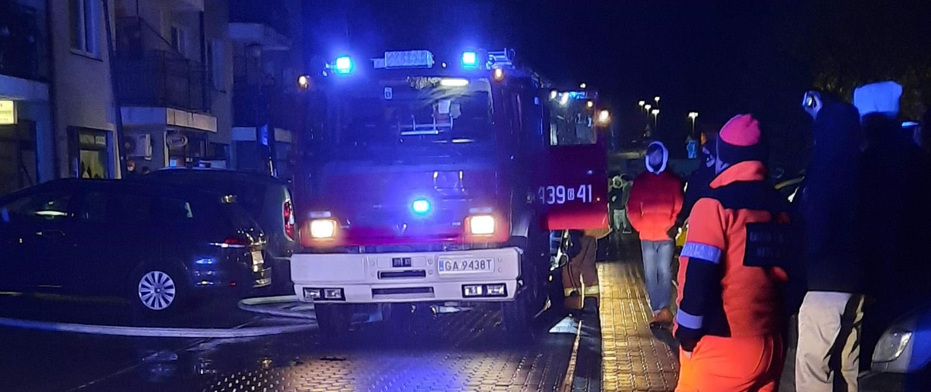 Jest ciemno. Wieczór. Czerwony wóz strażacki z włączonymi, niebieskimi światłami błyskowymi, stoi na drodze wyłożonej kostką brukową.