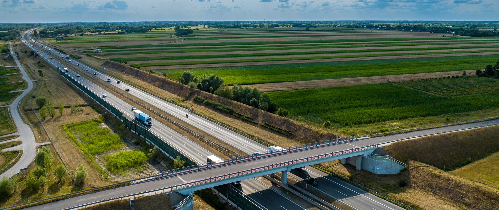 Zdjęcie lotnicze przedstawiające skrzyżowanie dróg ekspresowych na terenach rolniczych. Przez środek kadru biegnie dwujezdniowa droga z kilkoma pojazdami, nad którą przebiega wiadukt. Wokół widać rozległe pola uprawne i równinny krajobraz pod lekko zachmurzonym niebem.