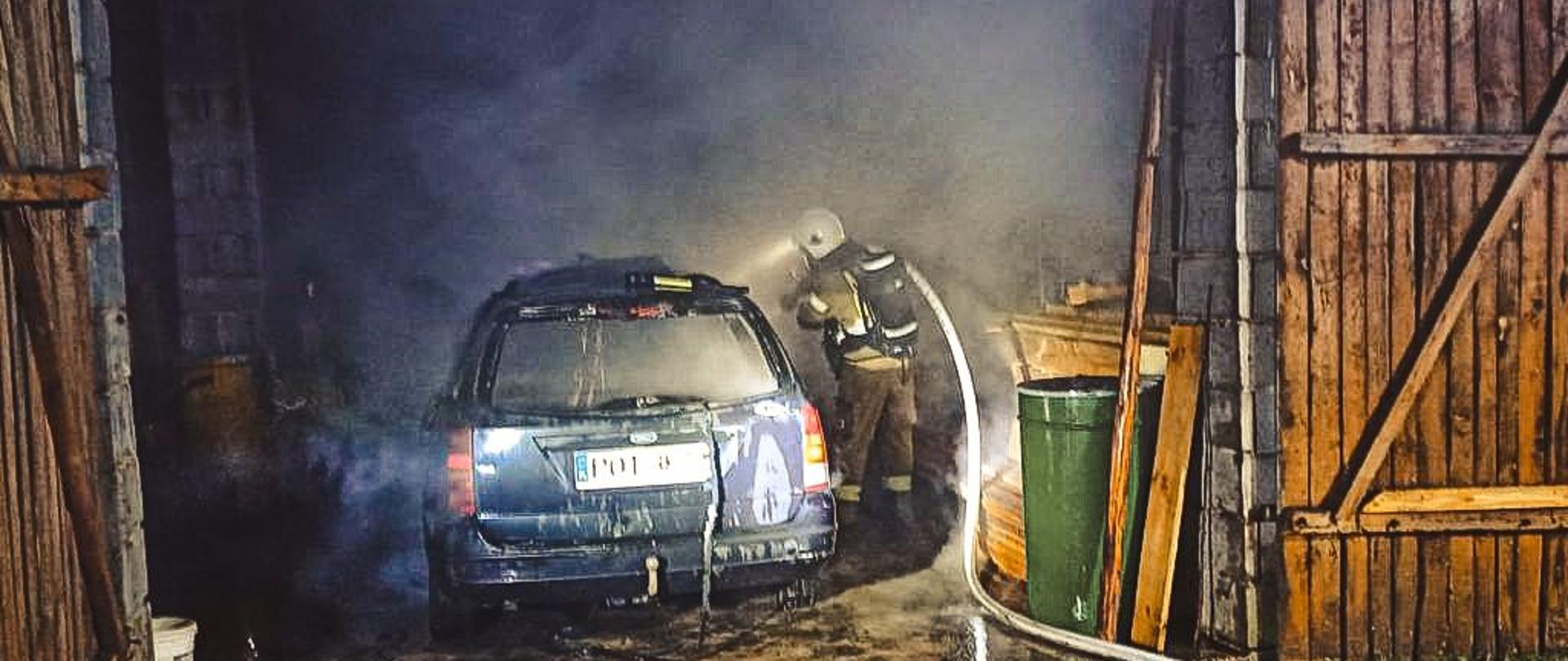 Samochód osobowy po pożarze stoi w budynku gospodarczym. Obok strażak gasi palący się pojazd.