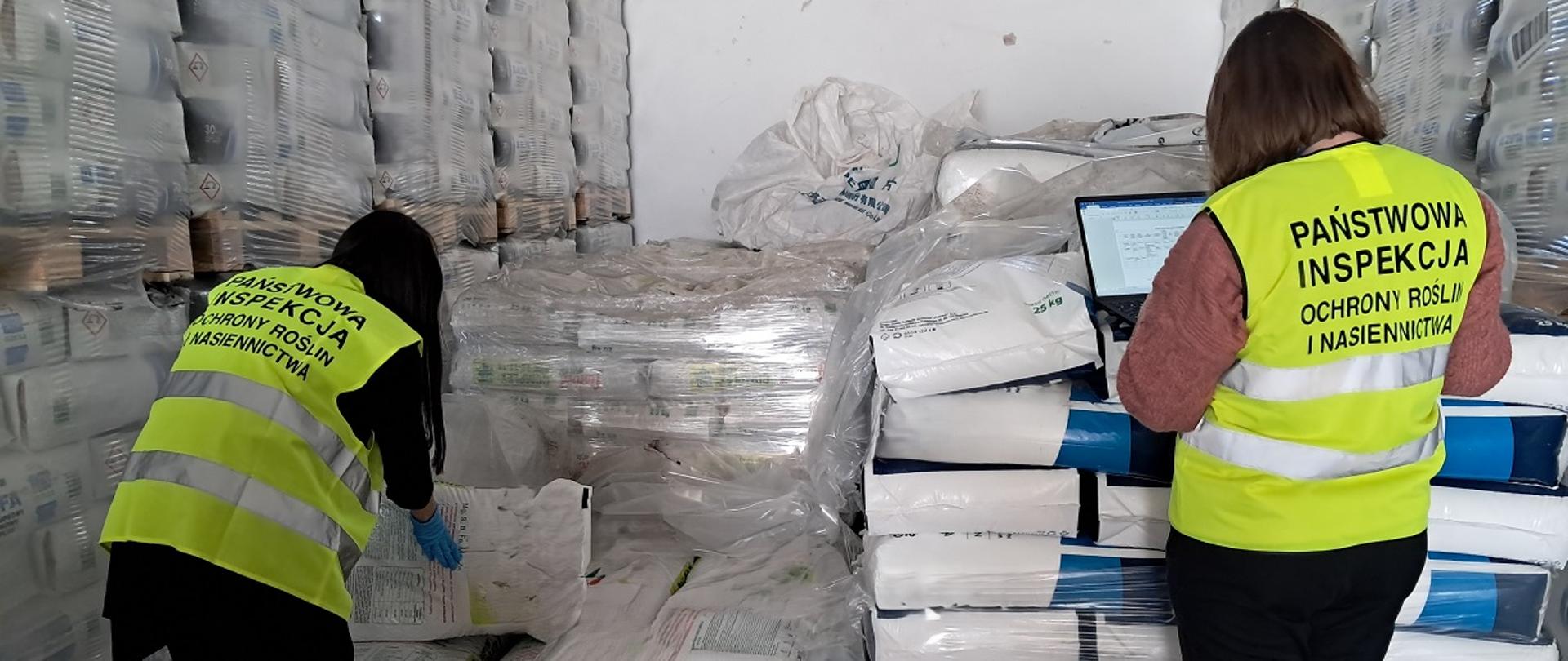 Dwóch inspektorów znajduje się w hali magazynowej. Ubrani są w żółte, odblaskowe kamizelki. W tle palety z nawozami. Jeden inspektor podnosi 25 kg worek nawozu, aby sprawdzić informacje zawarte na etykiecie. Drugi inspektor na laptopie zapisuje podyktowane przez niego dane. 