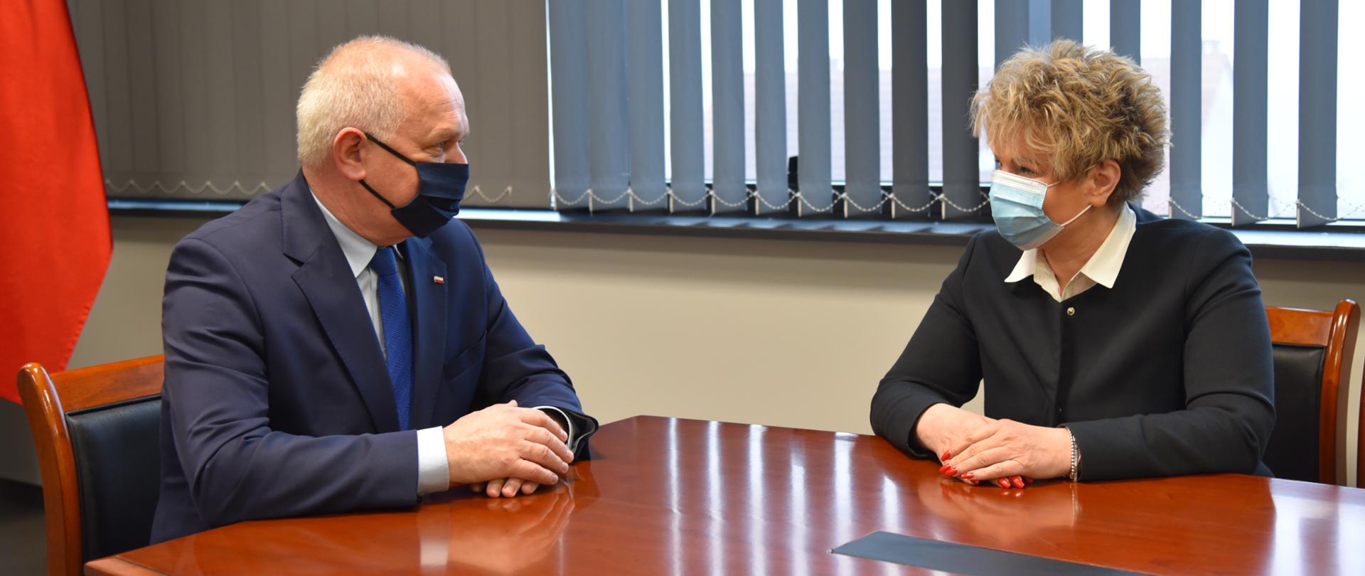 W tle okno z odsłoniętymi żaluzjami i kawałkiem biało-czerwonej flagi. Na pierwszym planie wojewoda rozmawia z burmistrz Małomic Małgorzatą Sendecką. Oboje mają założone maseczki. 