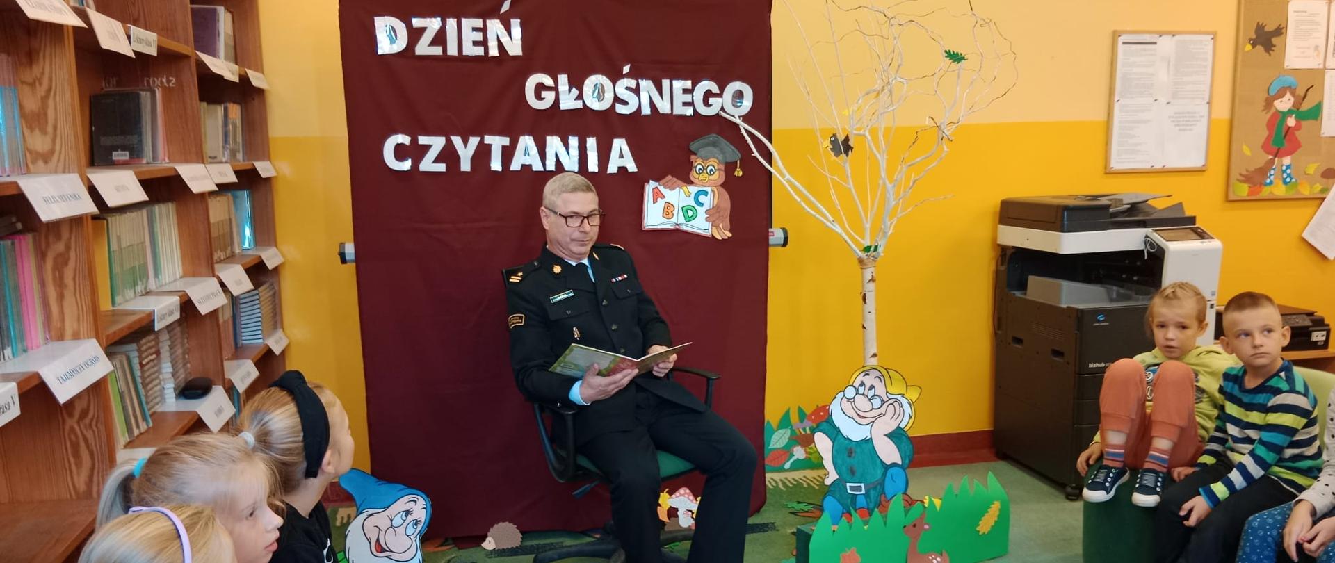 Na zdjęciu widać strażaka ubranego w mundur wyjściowy, trzyma książkę i czyta dzieciom.