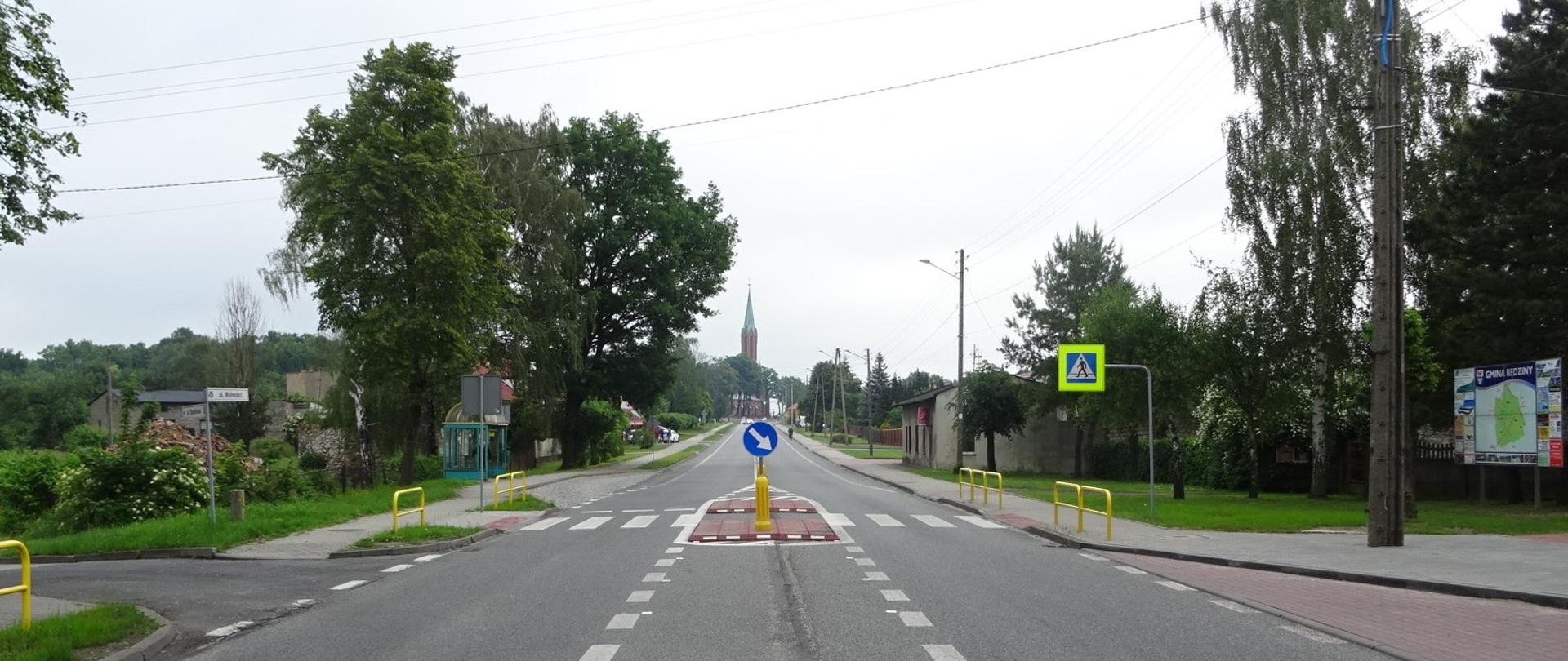 Przejście dla pieszych w ciągu DK91 w miejscowości Rędziny, na zdjęciu znajduje się przejście dla pieszych na drodze jednojezdniowej z azylem, w obrębie przejścia znajdują się przystanki autobusowe, w tle widać kościół