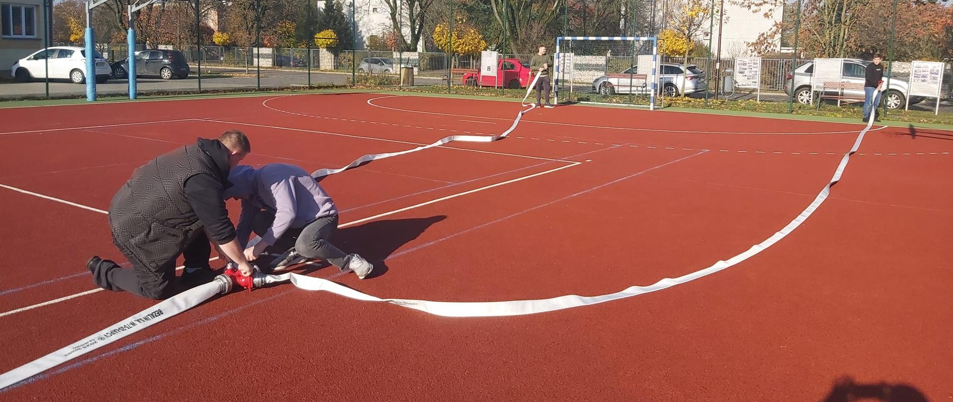Młodzież ćwiczy rozwijanie i skręcanie węża strażackiego na szkolnym boisku
