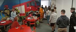 Zdjęcie zrobione na Sali Edukacyjnej. W tle czerwony wóz strażacki oraz strażacy gaszący pożar. Przy stolikach siedzą osoby wykonujące badanie. Za nimi tworzy się kolejka osób oczekujących na badanie. 