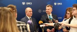 Minister stoi pod ścianką z napisem UMCS, obok niego stoi mężczyzna w garniturze który mówi do kilku mikrofonów trzymanych przez dziennikarzy.