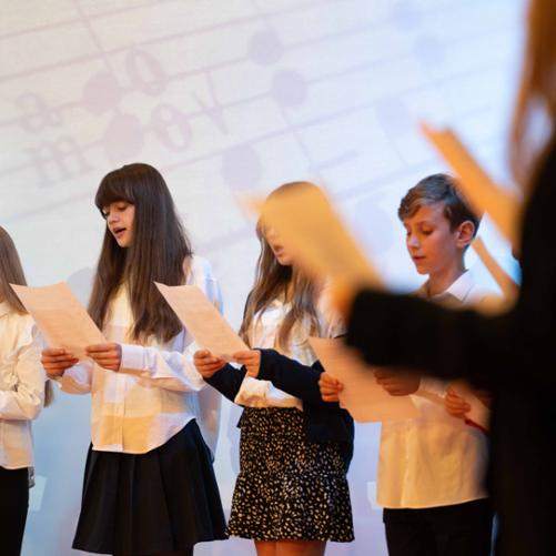 zdjęcie przedstawia członków chóru śpiewających na tle ekranu multimedialnego na scenie sali koncertowej