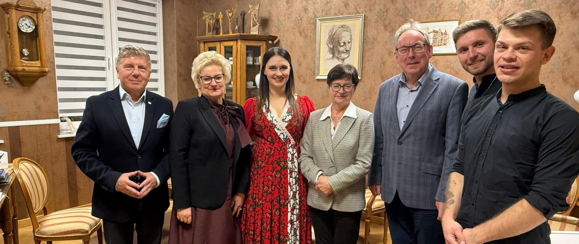 Grupa siedmiu osób stoi razem w eleganckim, przytulnie urządzonym pomieszczeniu. Wszyscy pozują do wspólnego zdjęcia i uśmiechają się w stronę aparatu. W centrum stoi kobieta w długiej czerwonej sukni, a po jej bokach znajdują się osoby w formalnych strojach, takich jak garnitury, marynarki i sukienki. W tle widać brązowe ściany z dekoracyjnymi obrazami, zegar ścienny oraz przeszkloną witrynę z figurkami.