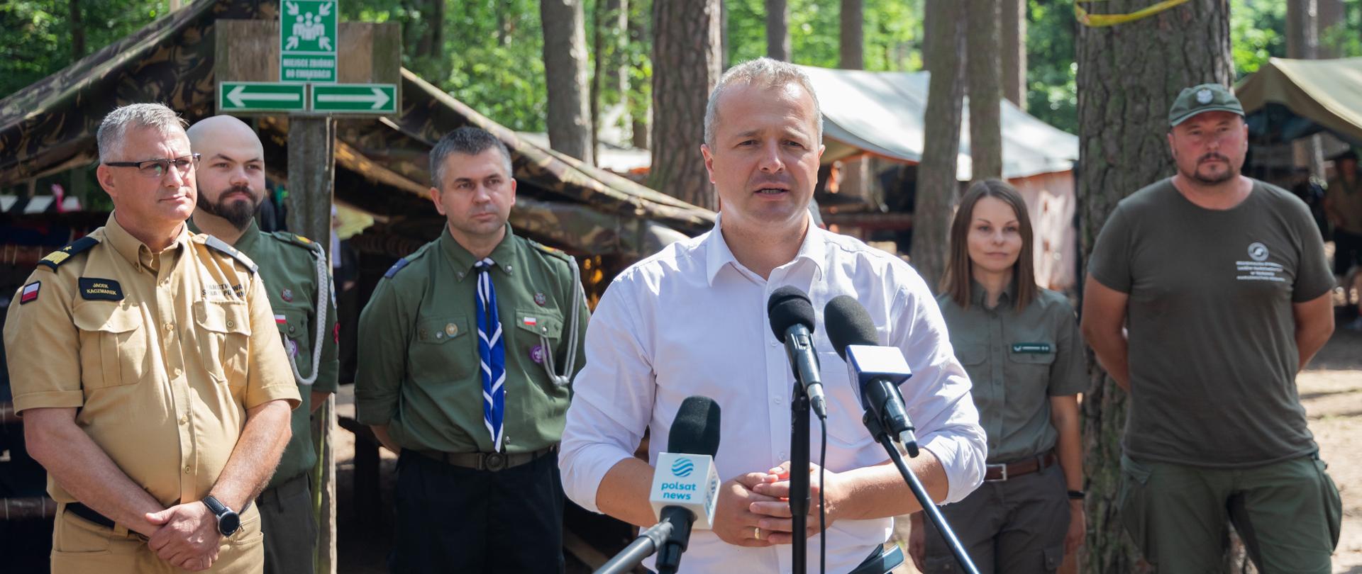 Jeden mężczyzna stojący przed mikrofonami i wypowiadający się. Za nim kilka osób