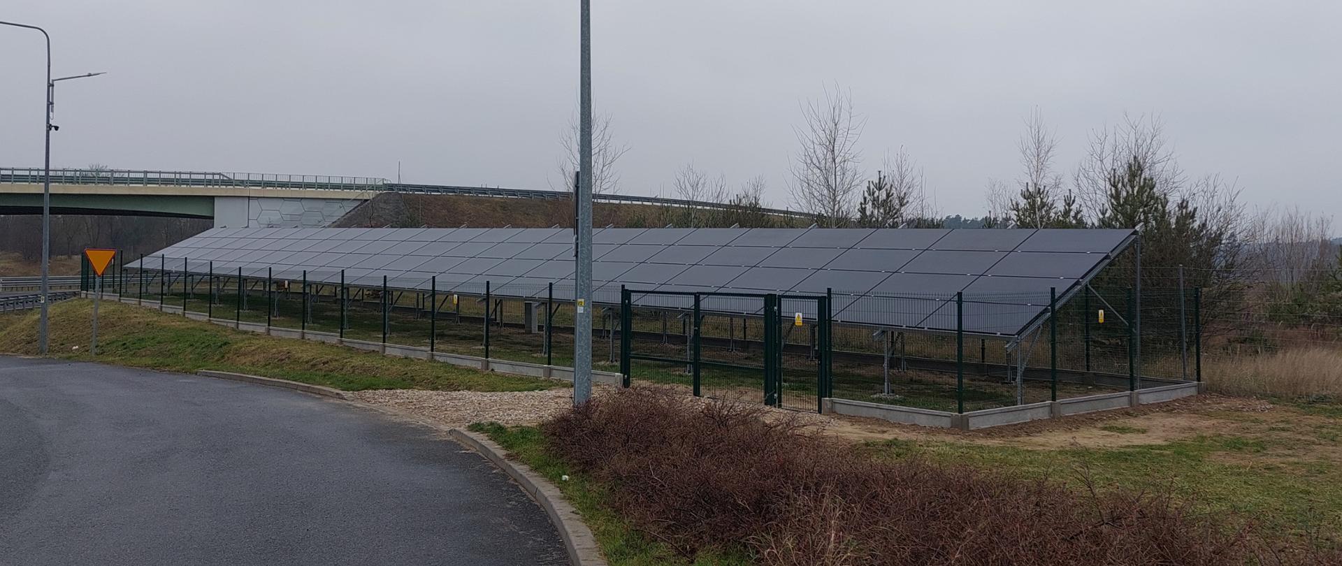 Na zdjęciu widać teren przy drodze, w pobliżu węzła lub zjazdu z autostrady lub drogi ekspresowej. Na pierwszym planie znajduje się asfaltowa jezdnia z krawężnikiem i fragmentem niskich krzewów po prawej stronie. W centralnej części obrazu stoi wysoki słup oświetleniowy. Za nim, ogrodzony metalowym płotem, znajduje się rząd paneli fotowoltaicznych ustawionych pod kątem, skierowanych w stronę nieba. Ogrodzenie jest wykonane z siatki na metalowych słupkach, a w jego obrębie widać konstrukcję wspierającą panele. W tle po lewej stronie widoczny jest nasyp z barierą energochłonną i fragmentem drogi, prawdopodobnie wiaduktu. W oddali rosną niewielkie drzewa i krzewy. Niebo jest całkowicie zachmurzone, co nadaje zdjęciu szary, zimowy charakter