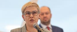 Wiceminister Mrówczyńska stoi i mówi do mikrofonu na stojaku.