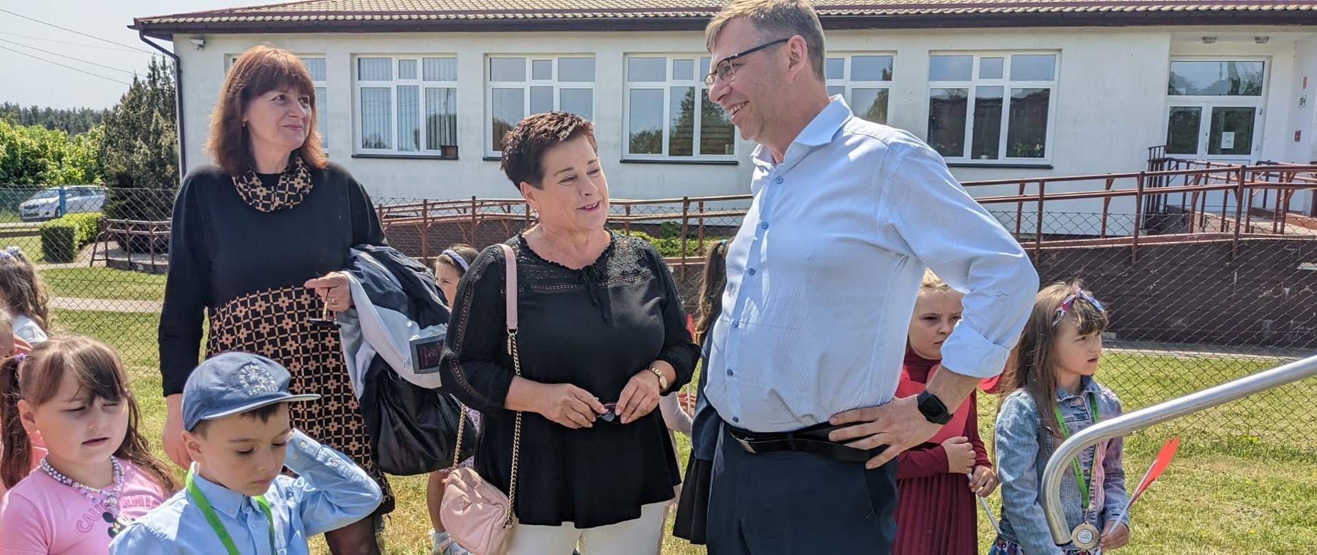 Uśmiechnięta Pani w czarnej koszulce rozmawia z Wojewodą, który również się uśmiecha. Na pierwszym planie i w tle zdjęcia są dzieci zaaferowane zabawą 
