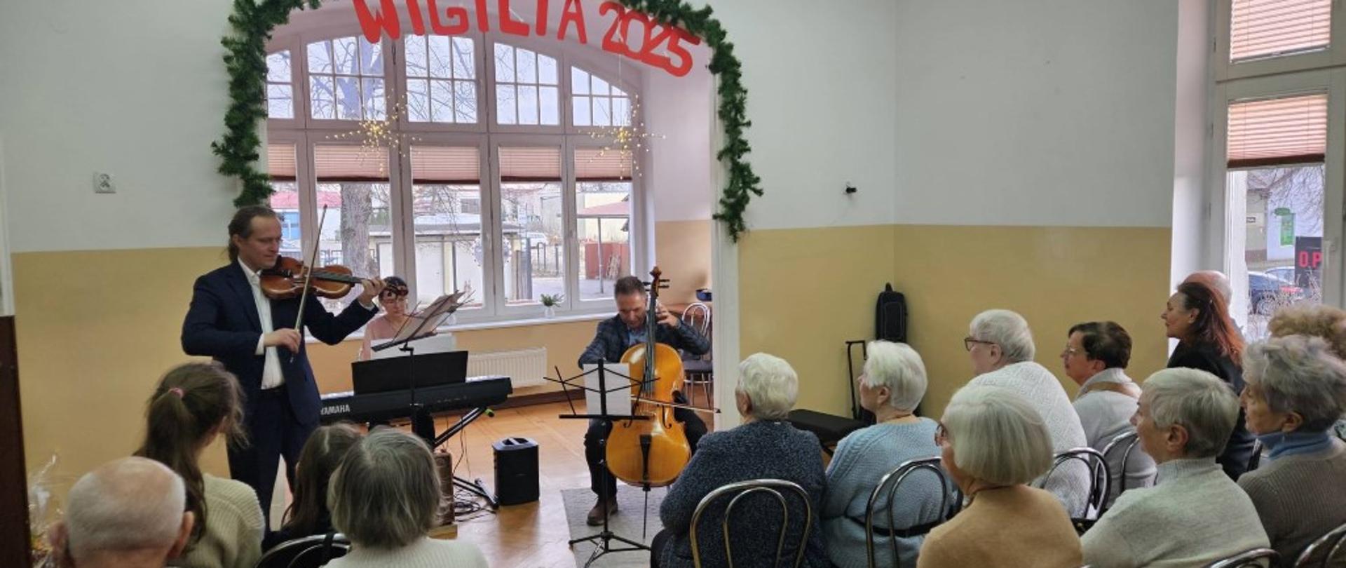 Zdjęcie kolorowe. Trójka muzyków grająca koncert dla publiczności w jasnej sali. Po lewej stronie mężczyzna w ciemnym garniturze grający na skrzypcach. W głębi kobieta siedząca przy czarnym pianinie cyfrowym marki Yamaha. Pośrodku mężczyzna grający na wiolonczeli, siedzący przodem do słuchaczy. Nad oknem w tle dekoracja z igliwia oraz czerwony napis "WIGILIA 2025". Publiczność siedząca na krzesłach tyłem i bokiem do obiektywu, składająca się z kobiet i mężczyzn, ubranych w swetry i żakiety. W tle duże okna z widokiem na zewnątrz, udekorowane świecącymi gwiazdami.