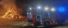 Nocna akcja straży pożarnej – wóz strażacki z włączonymi światłami stoi na polu, obok płonie duży stos siana. Z wozu prowadzi wąż gaśniczy, ogień oświetla otoczenie, a w tle widać drzewa.