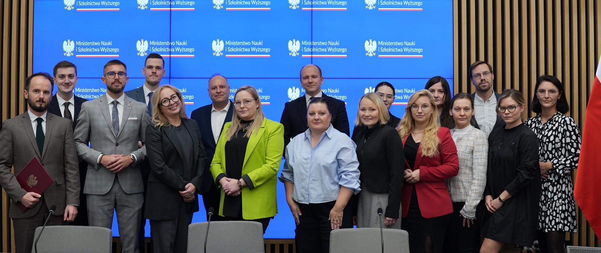 Zdjęcie zbiorowe, przed wbudowanym w ścianę z pionowych drewnianych listewek wielkim niebieskim ekranem stoi grupa ok. 20 osób, z prawej pod ścianą flagi Polski i UE.