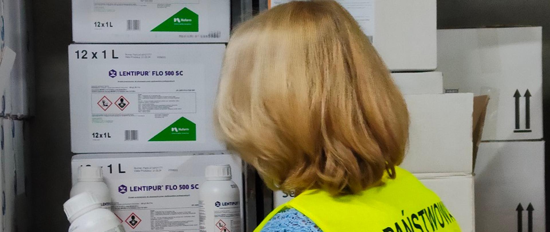 Inspektor znajduje się w hali magazynowej. Ubrany jest w żółtą odblaskową kamizelkę. Ogląda opakowanie środka ochrony roślin.