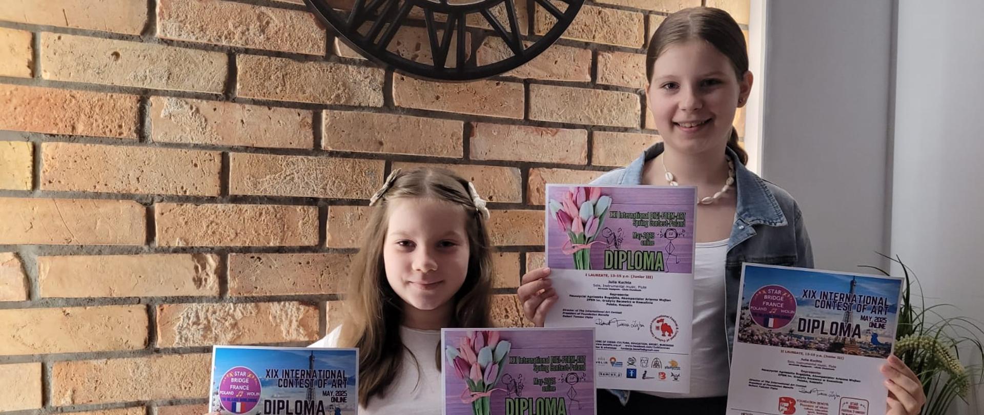 Uczennice ZPSM Maja i Julia z dyplomami na tle ceglanej ściany i zegara