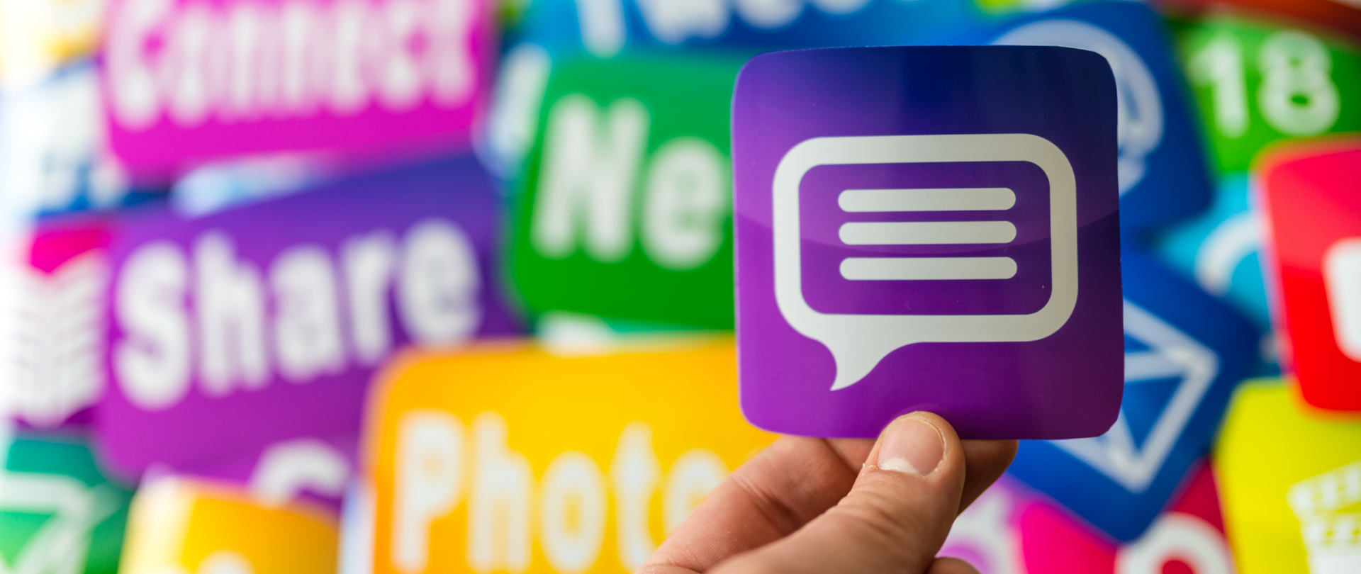 Na pierwszym planie dłoń trzymająca fioletowy obrazem z białą chmurką przypominającą symbol internetowego komentarza. W tle, rozmazane, kolorowe obrazki z symbolami poczty elektronicznej, tekstami share, photo, itp.