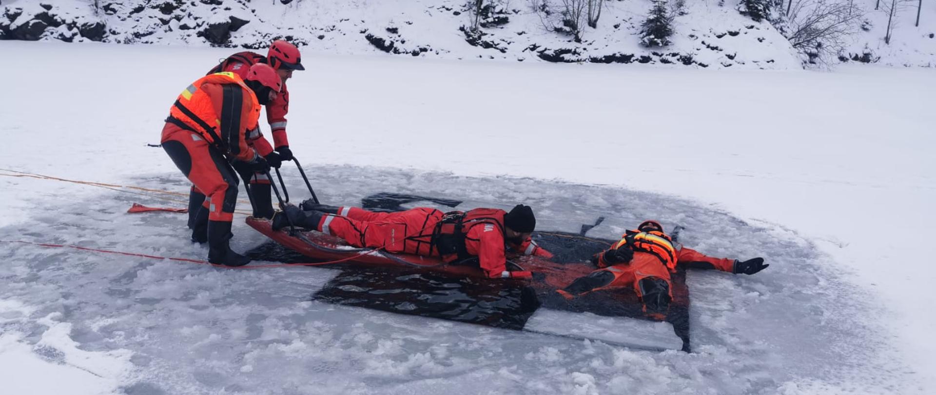 Na zdjęciu strażacy w pomarańczowych i czerwonych kombinezonach. Jeden strażak leży na plecach w wodzie, obok niego na saniach lodowych jest inny strażak, który go ratuje. Z lewej strony 2 strażaków stoi na lodzie i trzymają sanie lodowe.