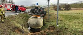 Przewrócona cysterna w rowie. Obok strażacy i pojazdy pożarnicze.